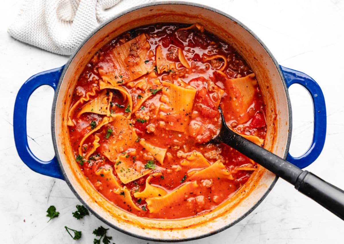 A blue Dutch oven with lasagna soup in it next to a cream knit cloth.