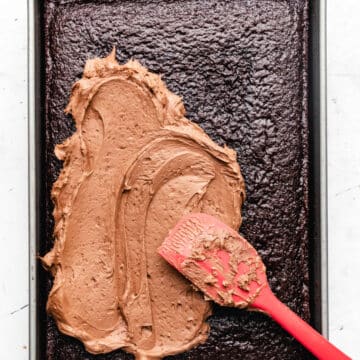 A red spatula spreading chocolate buttercream frosting on a chocolate sheet cake.