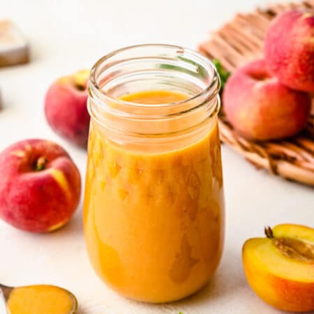 A glass jar filled with peach syrup next to fresh peaches.