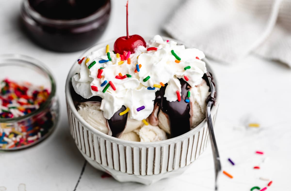 A hot fudge sundae next to dishes of hot fudge and sprinkles