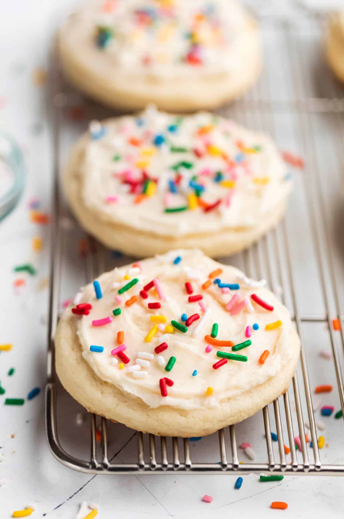 Three white frosted Lofthouse copycat cookies topped with rainbow sprinkles on a wire rack.