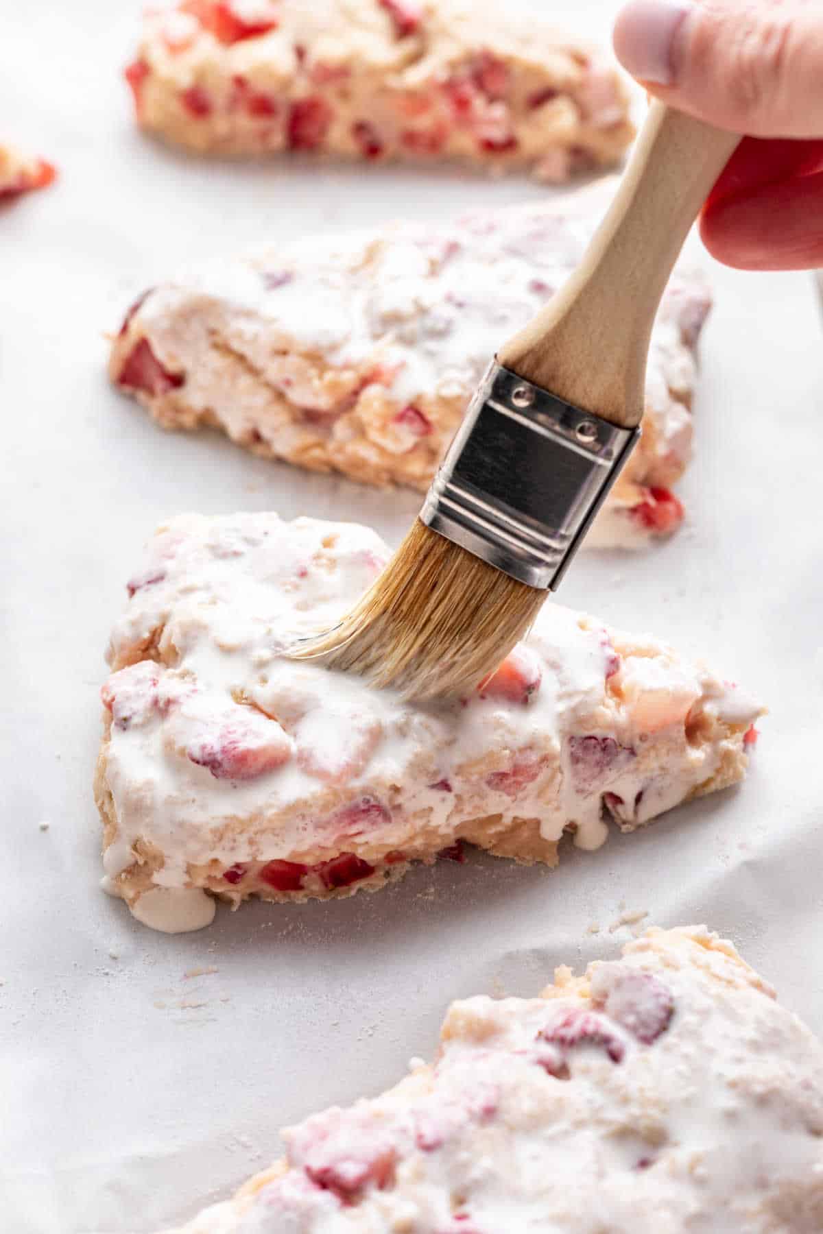 A pastry brush brushing cream onto an unbaked strawberry scone.