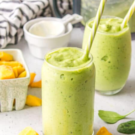 Two glasses of green smoothie with straws in them.