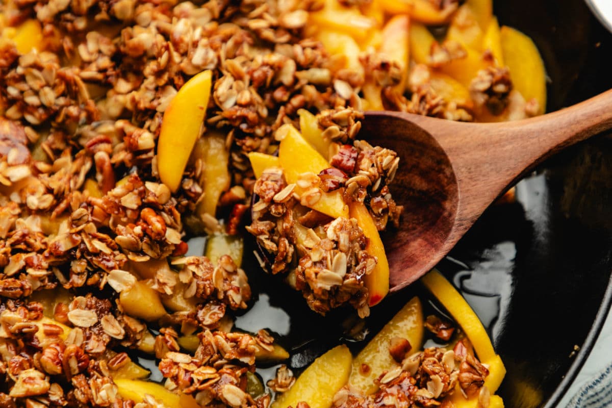 A wooden spoon scooping up peach crisp from a skillet. 