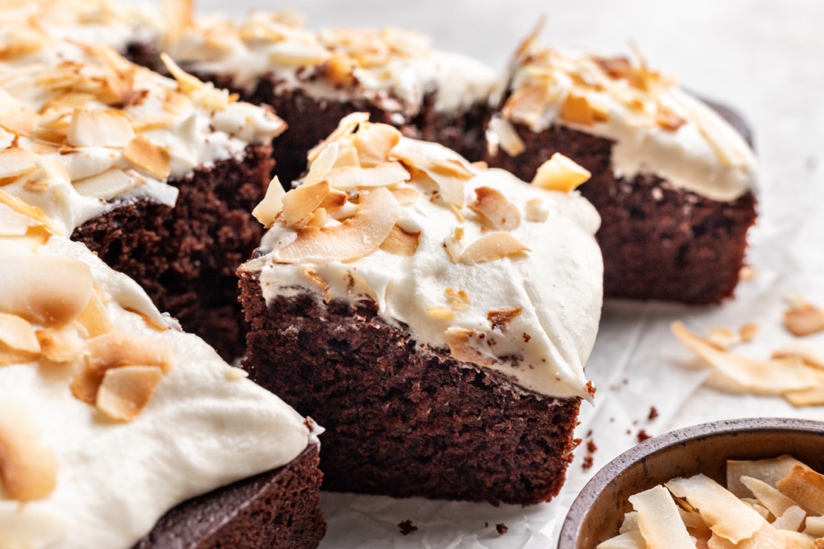 A cut piece of chocolate coconut cake next to a bowl of coconut chips.