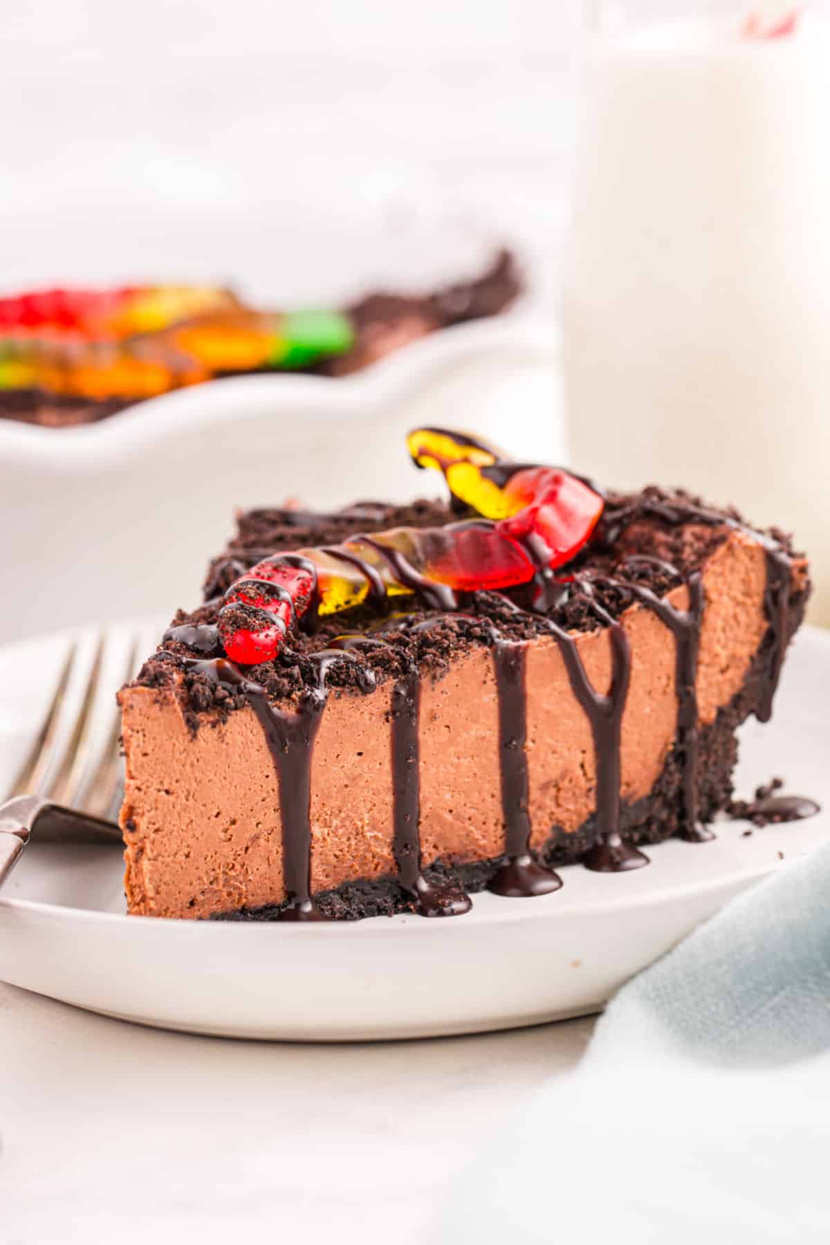 A slice of no bake dirt pie on a plate topped with chocolate syrup, crushed oreos, and a gummy worm.