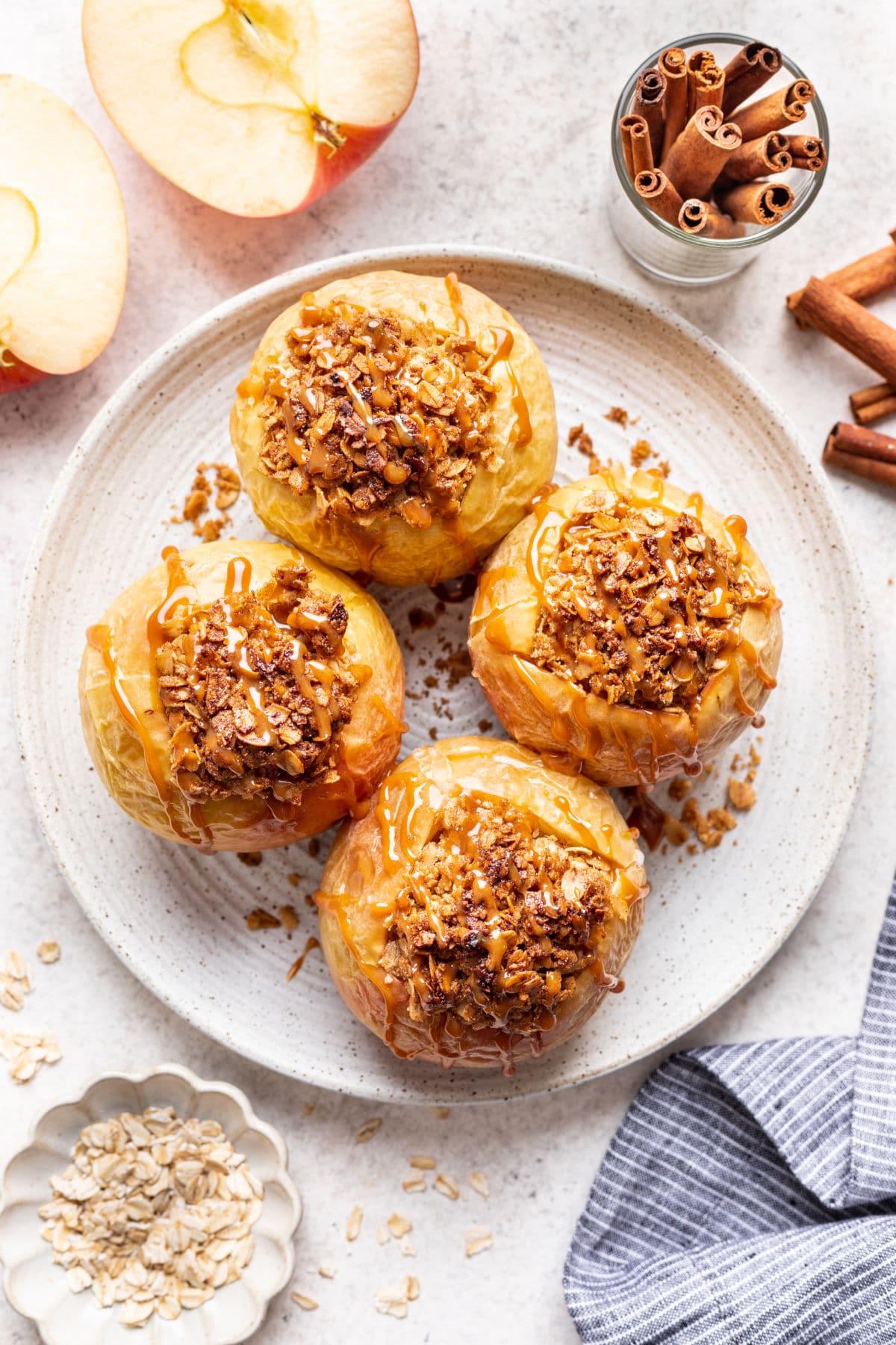 Four baked apples with oatmeal on a white plate next to cinnamon sticks. 