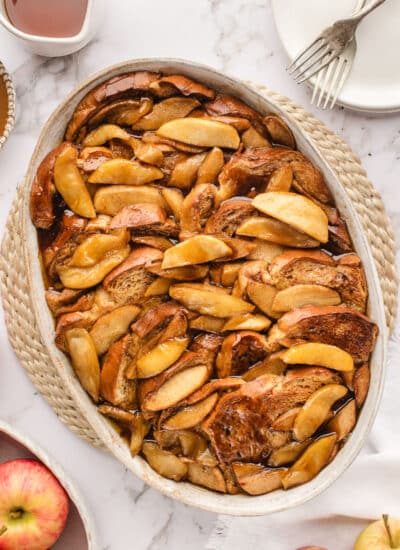 An oval baked filled with apple baked French toast next to a bowl of apples and syrup.