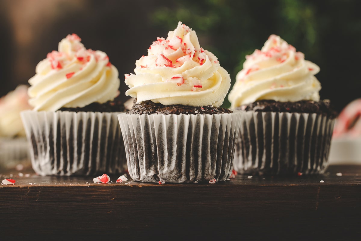 Three chocolate peppermint cupcakes in a row next to broken peppermint pieces. 