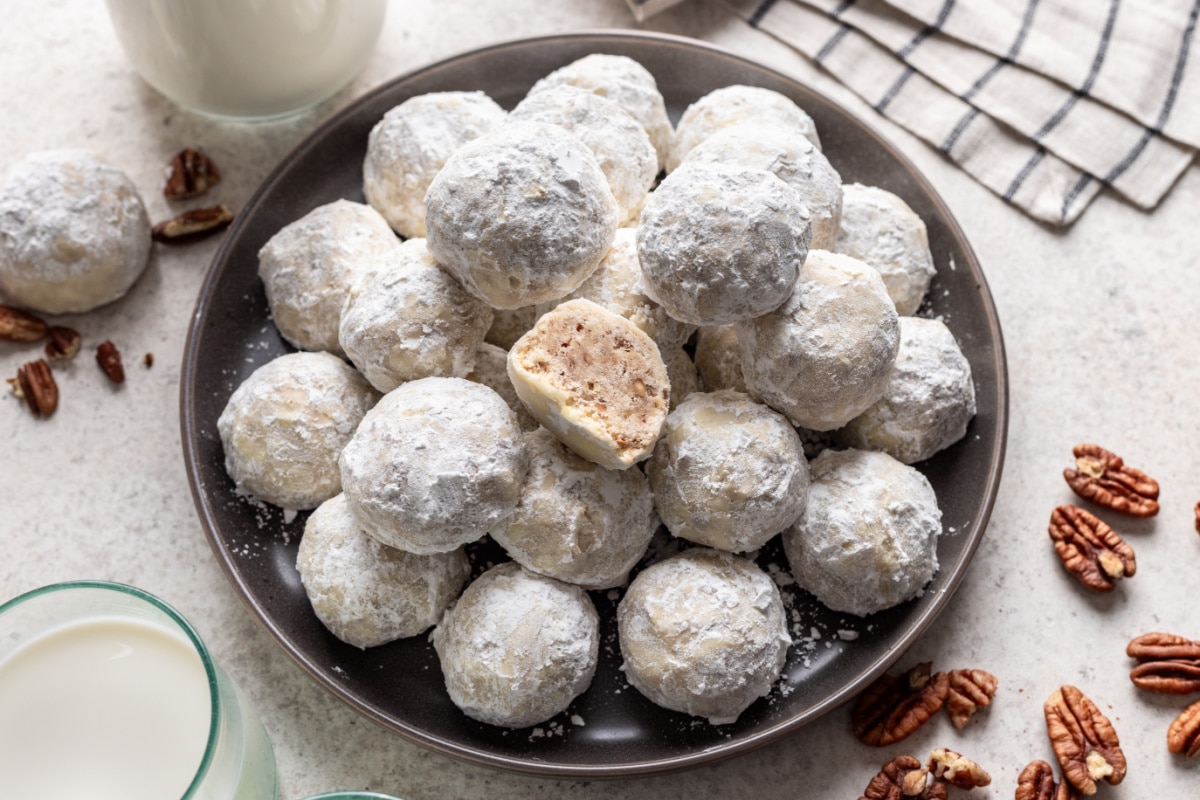 A plate of Mexican wedding cookies next to pecans and a striped kitchen linen.