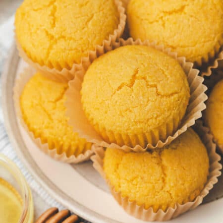 Honey cornbread muffins stacked on a plate next to a dish of honey.