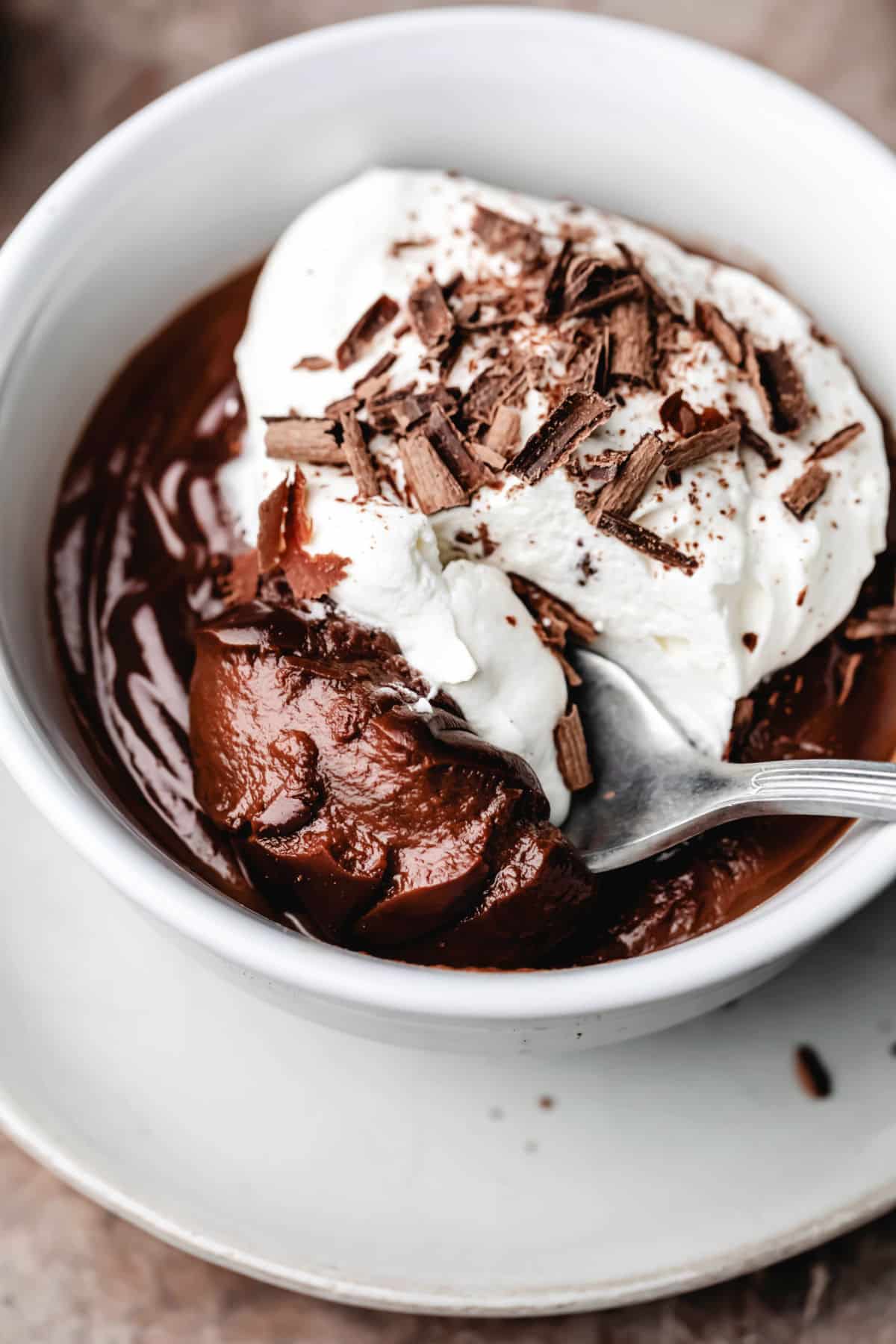 A spoon with a bite of chocolate pudding whipped cream and chocolate shavings in a dish of chocolate pudding.